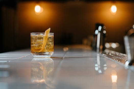 A stylish whiskey cocktail with zest garnish on a glossy bar counter in a dimly lit setting.